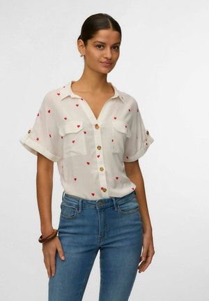 Femme portant une chemise blanche à manches courtes avec des motifs de cœurs rouges et un jean bleu, debout avec un bracelet marron au poignet.