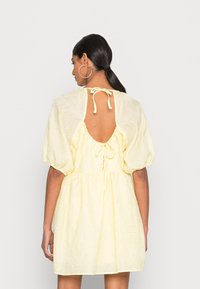 Vestido amarelo texturizado com mangas curtas bufantes, costas abertas e fecho com cordão no pescoço. Tecido leve com padrões subtis.