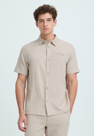 Homme aux cheveux bouclés portant une chemise beige à manches courtes boutonnée et un pantalon assorti, debout devant un fond clair uni.