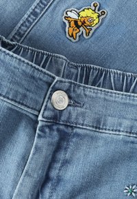 Nahaufnahme eines blauen Denimkleidungsstücks mit elastischem Bund, einem Metallknopf und einem bunten, bestickten Bienen-Patch.