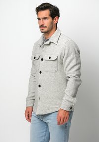 Grau strukturiertes Hemd-Jacke mit Kragen, schwarzen Knöpfen und zwei Brusttaschen, getragen über einem grauen Oberteil und kombiniert mit hellblauen Jeans.