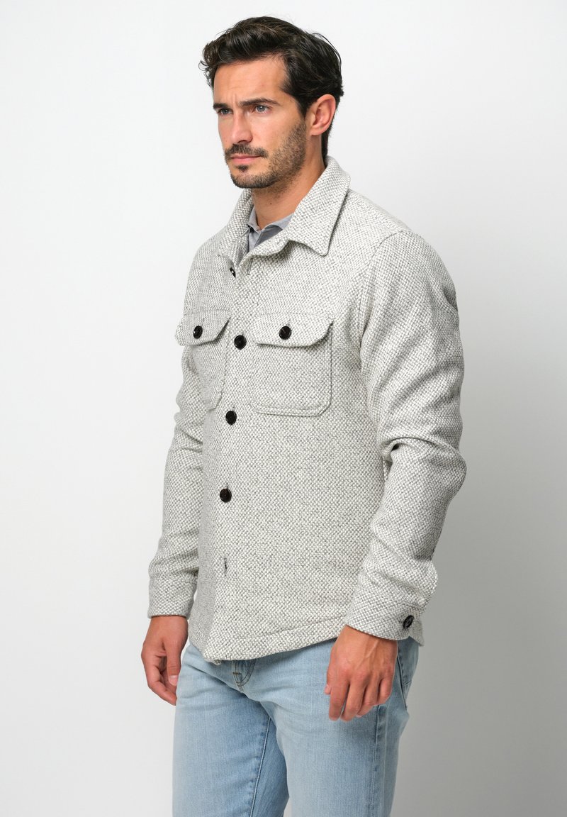 Grau strukturiertes Hemd-Jacke mit Kragen, schwarzen Knöpfen und zwei Brusttaschen, getragen über einem grauen Oberteil und kombiniert mit hellblauen Jeans.