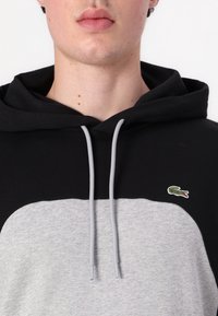 Sudadera con capucha negra y gris con un bolsillo tipo canguro en el frente, que presenta un pequeño logo de cocodrilo bordado y cordones grises.