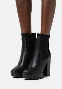 Bottes cheville en cuir noir avec un talon épais, des panneaux latéraux élastiques et une semelle extérieure en caoutchouc texturée avec un motif rainuré.