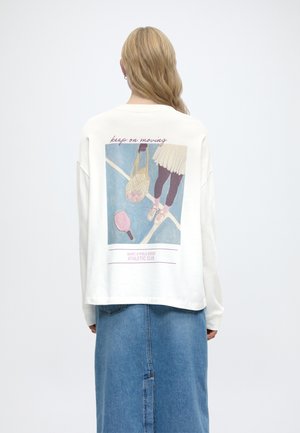 Άτομο με μακριά ξανθά μαλλιά που φορεί λευκό πουκάμισο με μακριά μανίκια, στο οποίο υπάρχει μια εικονογράφηση με θέμα το τένις και το κείμενο «keep on moving», καθώς και μπλε φούστα από τζιν.