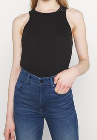 Top in maglia nera a costine con scollo alto, abbinato a jeans attillati blu scuro. La mano del modello è appoggiata nella tasca dei jeans.