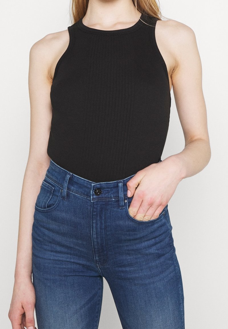 Top in maglia nera a costine con scollo alto, abbinato a jeans attillati blu scuro. La mano del modello è appoggiata nella tasca dei jeans.