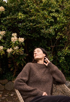 Femme aux cheveux foncés portant un pull marron, assise détendue dans un fauteuil en osier, les yeux fermés, près de buissons verts feuillus avec des fleurs blanches.