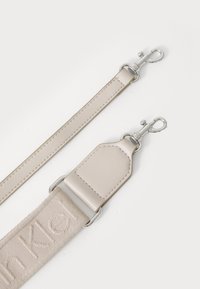 Beige verstellbarer Gurt mit geprägtem Logo, an beiden Enden mit silbernen Hardware-Clips und einem strukturierten Stoffabschnitt.