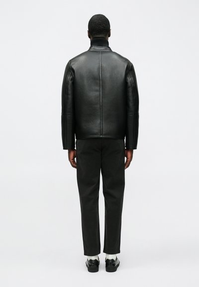 Schwarze Lederjacke mit hohem Kragen und strukturierten Schultern, kombiniert mit dunkler Hose und schwarzen Schuhen, aus der Rückansicht.