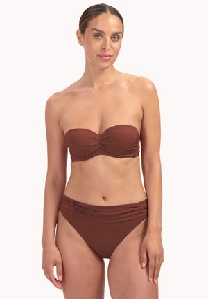 Femme portant un haut de bikini marron sans bretelles et un bas de bikini taille haute assorti, debout devant un fond clair uni.