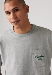 Pelēks īsām piedurknēm T-krekls, kas izgatavots no mīksta auduma, ar kreiso krūšu kabatu un zaļu izšuvumu "Levi Strauss & Co." logo.