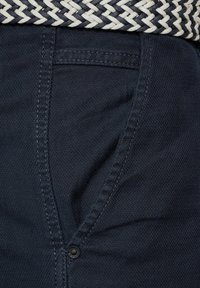 Marineblauwe broek met een gestructureerde stof, zichtbare naden, een voorzak en een knoopsluiting. Er is een geweven riem in zwart en wit aanwezig.