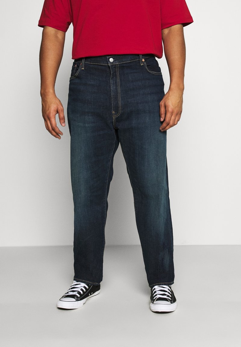 Homme portant un jean en denim bleu foncé, une chemise rouge et des baskets noires et blanches, debout devant un fond blanc.