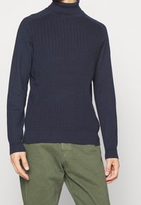 Manlig modell i en marinblå räfflad polo-tröja och olivgröna byxor, stående mot en enfärgad ljus bakgrund.