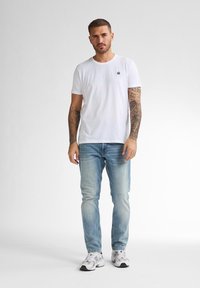 Camiseta de algodón blanca con un pequeño logo negro en el pecho, combinada con pantalones vaqueros de mezclilla azul claro y zapatillas blancas con acentos negros.