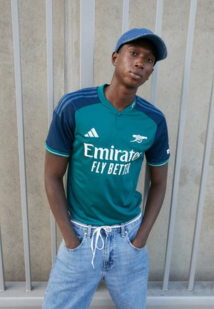 Maillot de sport à manches courtes vert et bleu marine avec texte blanc, jean en denim bleu, texture légère et casquette bleue. Les mains dans les poches.