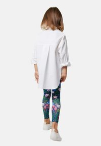 Biała, oversize koszula, legginsy w kwiatowy wzór w kolorze niebieskim, zielonym i różowym oraz białe buty typu slip-on noszone przez dziecko widziane z tyłu.