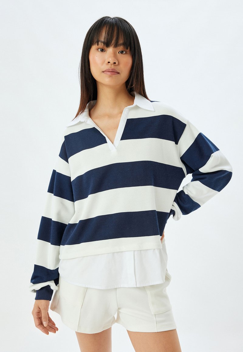 Maglione a righe blu navy e bianche con maniche lunghe, scollo a V e colletto bianco, in tessuto morbido, vestibilità comoda, indossato sopra una camicia bianca e pantaloni corti.