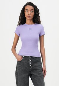 T-shirt lilas à côtes avec des manches courtes, arborant un petit logo sur la poitrine. Associé à un jean noir taille haute avec fermeture à bouton.