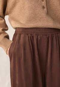 Braune Hosen mit elastischem Bund und glatter Textur, mit Seitentaschen und weiter Passform, kombiniert mit einem beigefarbenen Strickpullover.