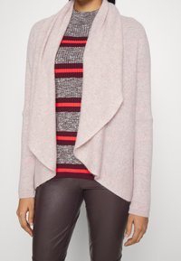 Hellrosa offene Strickjacke aus weichem Material mit langen Ärmeln und einem drapierten Kragen, getragen über einem gestreiften Strickoberteil in Grau und Rot.