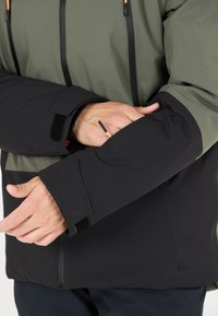 Vandtæt jakke med en olivengrøn overdel og sort underdel, med Velcro håndledslukninger og en subtil tekstur.