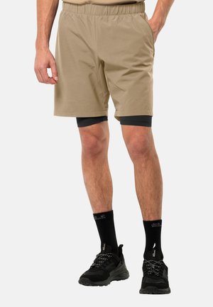 Beige Shorts mit elastischem Bund, ausgestattet mit Seitentaschen und einem lockeren Schnitt. Getragen über schwarzen Kompressionsshorts und kombiniert mit schwarzen Sneakers.