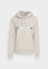 Beige mikina s kapucí a přední kapsou, s šňůrkami a potiskem "Hollister California" v metalické stříbrné barvě, vyrobená z měkké bavlněné směsi.