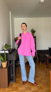 Femme portant une chemise boutonnée rose vif, un jean bleu clair, des talons pointus marron, tenant un sac tissé foncé dans un salon avec des plantes.