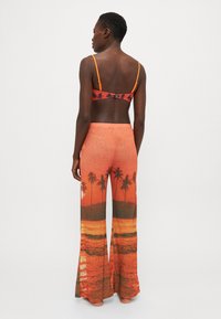 Bralette naranja con patrón de palmeras, combinado con pantalones de pierna ancha que presentan una escena de playa al atardecer en tonos de color similares, tejido suave.