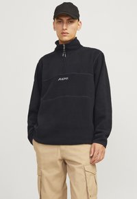 Pull en polaire noir avec un col montant, une fermeture éclair sur le devant à mi-hauteur, et un logo. Présente une texture et une couleur unie. Porté avec un pantalon cargo beige.