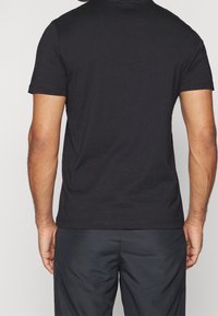 Černé tričko s krátkým rukávem zobrazené zezadu, s hladkým materiálem a uvolněným střihem. Minimalistický design, bez vzorů a doplňků.