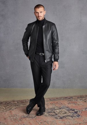 Chaqueta bomber negra de cuero, ajustada, con cierre de cremallera y cuello alto. Llevada sobre un cuello de tortuga negro y combinada con pantalones negros y botas.