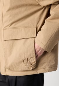 Šviesiai ruda striukė su laisvu siluetu, turinti didelę priekinę kišenę ir siuvinėtą "CK" logotipą ant apatinio krašto. Sklandi tekstūra ir minimalus dizainas.