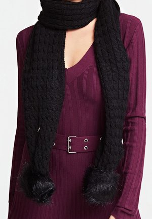 Femme portant une robe en maille côtelée violette avec une ceinture assortie et une longue écharpe noire tricotée avec des pompons en fausse fourrure aux deux extrémités.