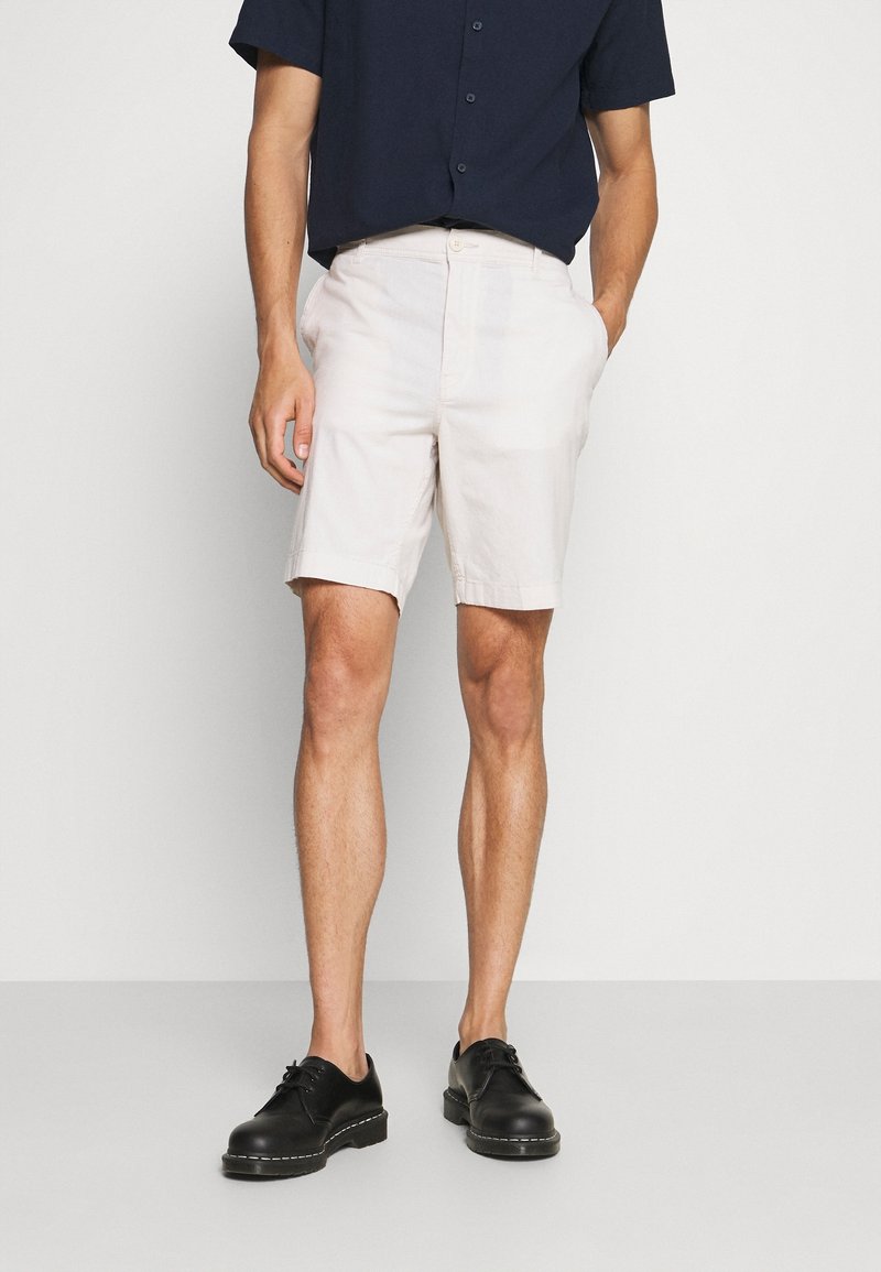 Bermudas en coton de couleur claire avec poches latérales, ourlets retroussés et fermeture à bouton, associées à une chemise à manches courtes sombre et des chaussures noires.