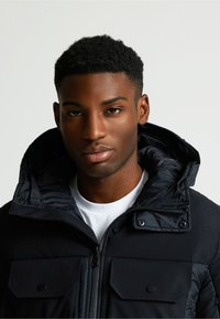 Čierna bundu v štýle pufáku s vysokým golierom, veľkými prsiami, predným zipsom a lesklou textúrou. Nosená cez biele tričko.