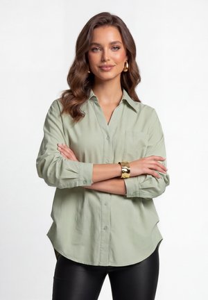 Jonge vrouw met golvend bruin haar die een lichtgroen blouse met knopen draagt, zwarte broek, gouden creooloorbellen en een gouden armband, armen gekruist.