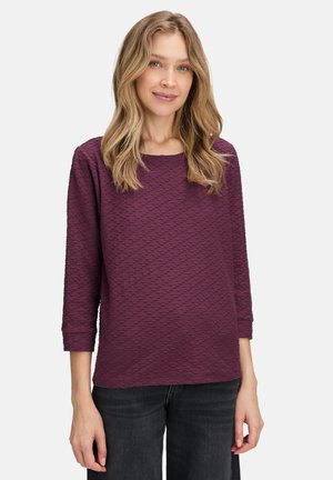 Camisetta viola con texture, colletto rotondo e maniche a tre quarti, caratterizzata da un motivo geometrico. Abbinata a jeans scuri.