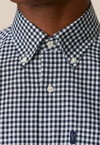 Chemise à carreaux bleu marine et blanche avec un col classique, dotée de boutons blancs et d'un petit logo en bas à gauche.