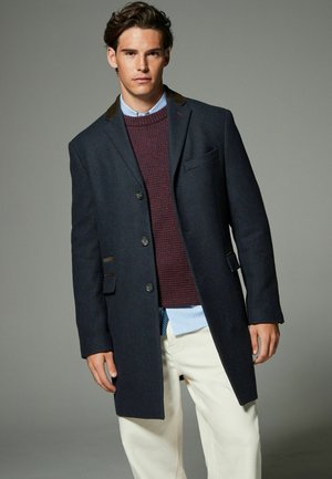 Marineblauer Wollmantel mit einreihigem Design, fünf Knöpfen und einer Brusttasche. Kombiniert mit einem kastanienbraunen Strickpullover und einem hellblauen Hemd.