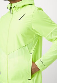 Giacca Nike verde neon con cappuccio, chiusura zip frontale e tasche laterali. Testo "STORM-FIT" sul polsino; design elegante, leggero e resistente all'acqua.