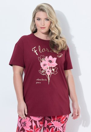 Vrouw met lang golvend blond haar draagt een bordeaux t-shirt met roze bloemen en de tekst "Floral where beauty grows" en broeken met een bloemenpatroon.