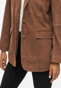 Ruskea mokkanahkablazeri, jossa on kaksi nappia ja läppätaskut, käytetty valkoisen paidan ja mustien housujen päällä, kädet näkyvät kyljissä.