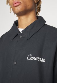 Jeune personne avec des dreadlocks clairs portant une veste noire à boutons avec le texte blanc « Converse » sur la poitrine, par-dessus une chemise blanche.