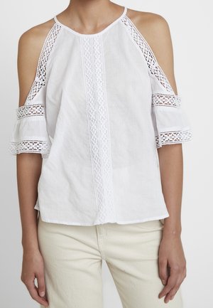 Kvinde iført en hvid cold-shoulder top med blonde detaljer og flæseærmer, parret med lyse beige bukser.