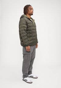 Gepolsterte olivgrüne Jacke mit Kapuze, graue Cargo-Hosen und weiße Schuhe mit schwarzen Streifen, stehen vor einem weißen Hintergrund.