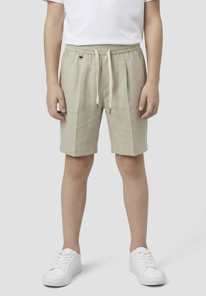 Garçon portant un short beige avec cordon, des baskets blanches et une chemise blanche à manches courtes, debout devant un fond clair uni.