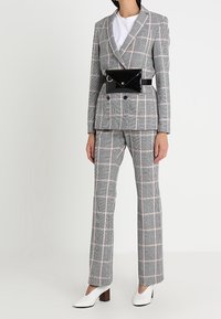 Vrouw in een grijs geruit dubbelzijdig pak met roze lijnen, witte top, zwarte laarzen met glanzend leer, en witte blokhak schoenen.